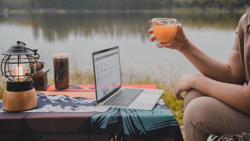 A person holding a glass of orange juice sits beside a table with a lit lantern, a laptop displaying a calendar, and a jar near a lake.